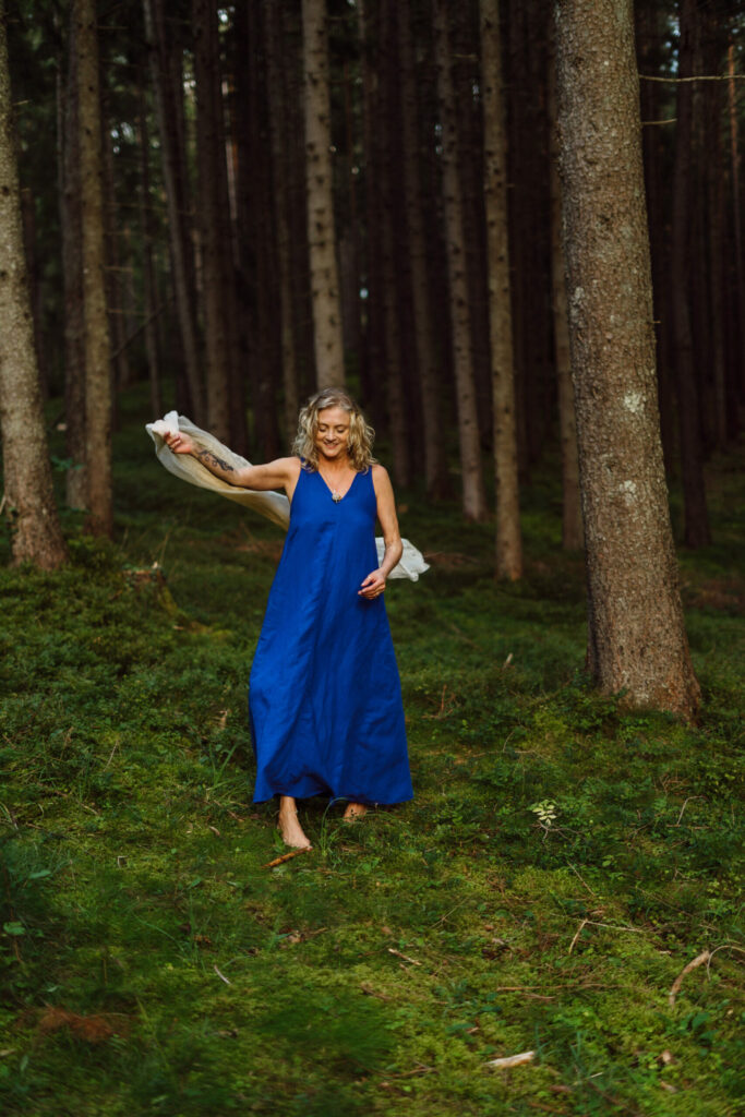 Heike Wangler tanzt mit weißem Tuch in einem blauen Kleid schwungvoll zwischen Fichtenbäumen.