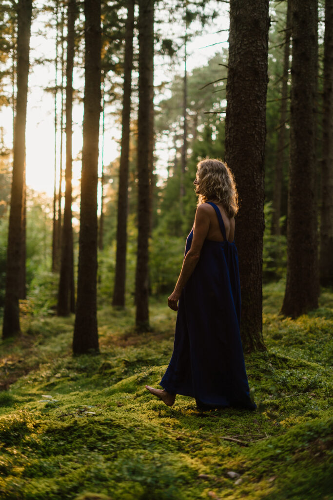 Heike Wangler wandert barfuß in einem blauem Kleid über moosbewachsenen Waldboden im Fichtenwald.